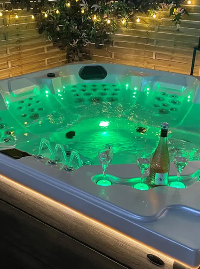 Where Comfort Meets Craftsmanship: Our Hot Tub Tale 1 A hot tub with green LED lights, bubbling water, and built-in cup holders. Three wine glasses and a bottle of sparkling wine rest on the tub’s edge. Wooden fencing and fairy lights create a cosy backdrop.