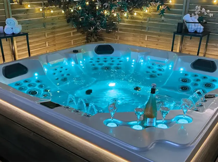 A large, illuminated jacuzzi with bubbling water sits in a cosy outdoor space. It is surrounded by wooden fencing, fairy lights, drinks, and towels, creating a relaxing, inviting atmosphere reminiscent of Dubai’s luxury service.