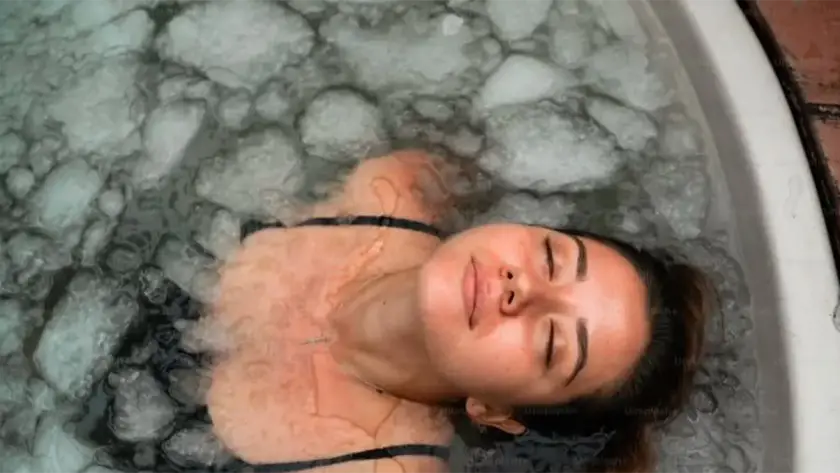 A woman with closed eyes relaxes in a tub filled with ice and water, wearing a black swimming costume. Her face appears calm as she leans back in the icy bath.