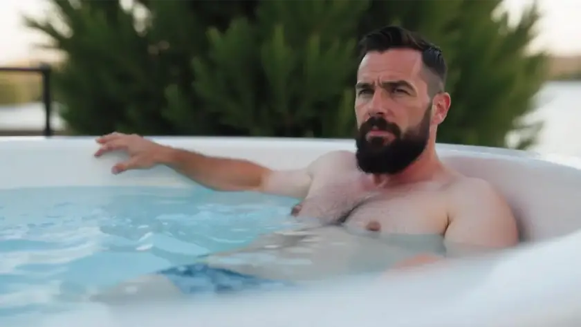 A man with a beard relaxes in an outdoor hot tub, resting one arm on the edge. Green trees and water create a peaceful backdrop behind him, capturing a perfect jacuzzi moment.