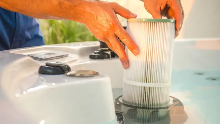 Close-up of hands performing hot tub maintenance by installing or removing a cylindrical pleated filter from the water, with control knobs and part of the hot tub visible in the foreground—ideal for spa care in the UAE.