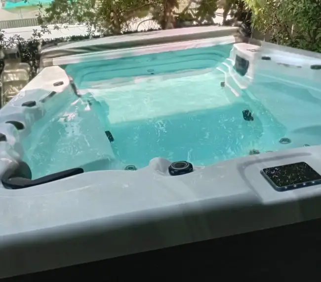 A clean, empty hot tub filled with clear blue water sits outdoors in a lush garden, surrounded by vibrant foliage and trees, with sunlight reflecting on the water’s surface.