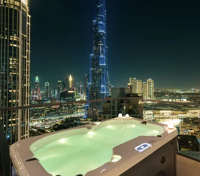 A rooftop hot tub glows at night atop a luxury flat, with city lights and skyscrapers in the background, including the illuminated Burj Khalifa towering over the Dubai skyline.
