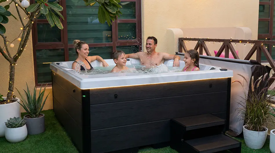 TubLease Hot Tub Rental and Sales 6 Four people, two adults and two children, relax and smile together in a hot tub jacuzzi surrounded by plants and fairy lights in a cosy outdoor patio setting.