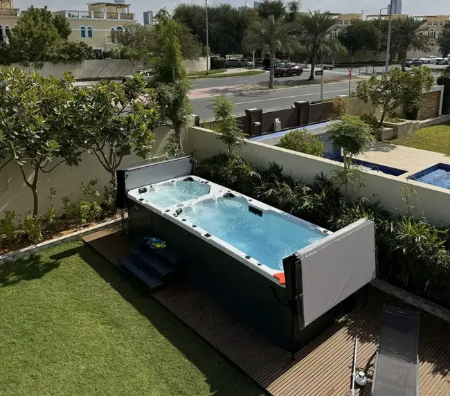 A rectangular swim spa sits on a wooden decking in a green garden, surrounded by lush plants and trees, offering a perfect spot for exercise with views of a residential street and houses in the background.
