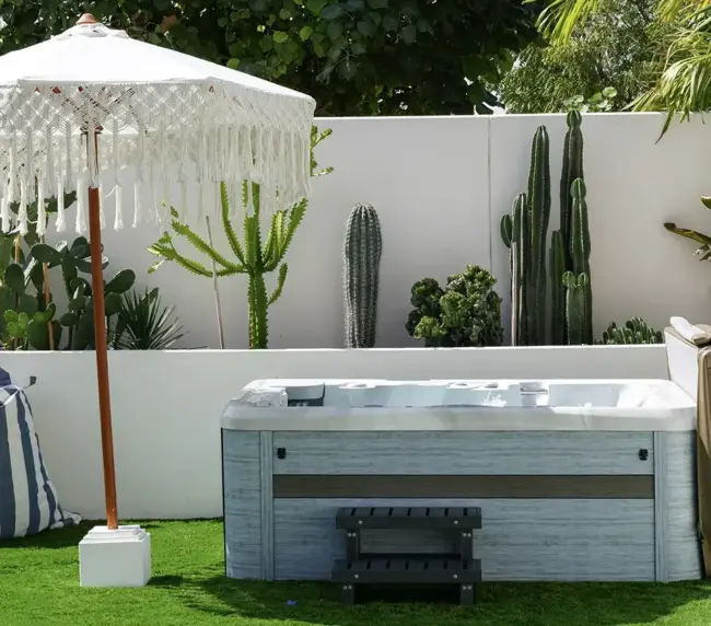 A modern jacuzzi sits on artificial grass beside a fringed white parasol, with tall cacti and leafy green plants creating a lush garden landscape against a sunny white wall.
