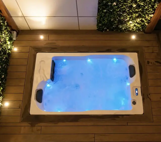 Overhead view of a rectangular hot tub with blue LED lights, bubbling water, and two headrests, set on a wooden decking balcony with greenery on two sides and small recessed lights along the edges—perfect for a relaxing rental stay.