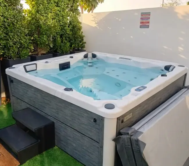 A clean, modern outdoor jacuzzi filled with water sits on artificial grass beside a white wall and tall green plants, creating a serene landscape. A folded hot tub cover and black steps are positioned next to it in Abu Dhabi.