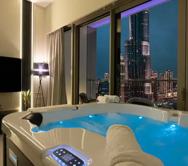 A modern city centre hotel room with a lit jacuzzi facing floor-to-ceiling windows, offering a night view of illuminated skyscrapers. Rolled towels rest on the jacuzzi’s edge as city lights sparkle outside, evoking a luxury flat feel.